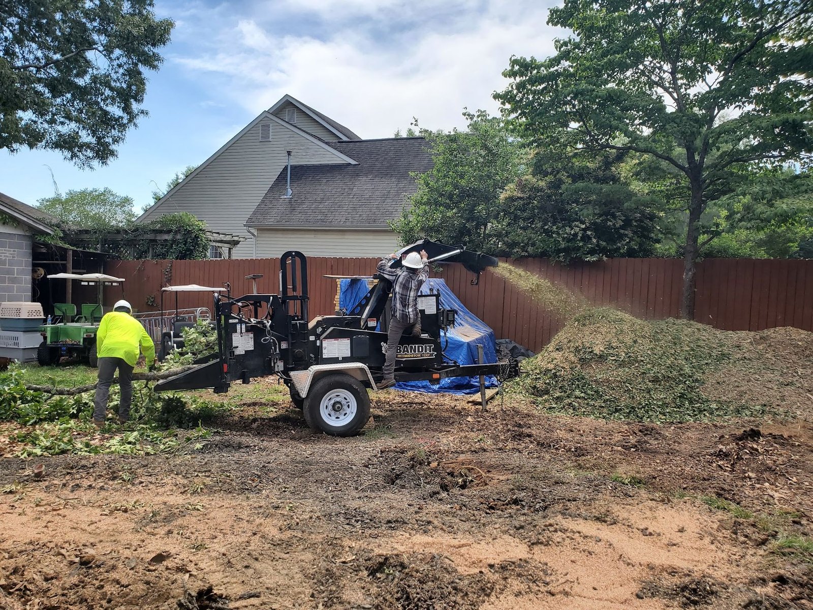 Crew member feeding brush into the chipper beside a house with a large mound of fresh chips on the ground