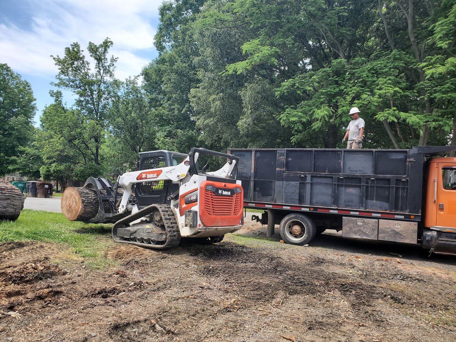 Skid steer loading a heavy log into the bed of a dump truck with a crew member on the truck