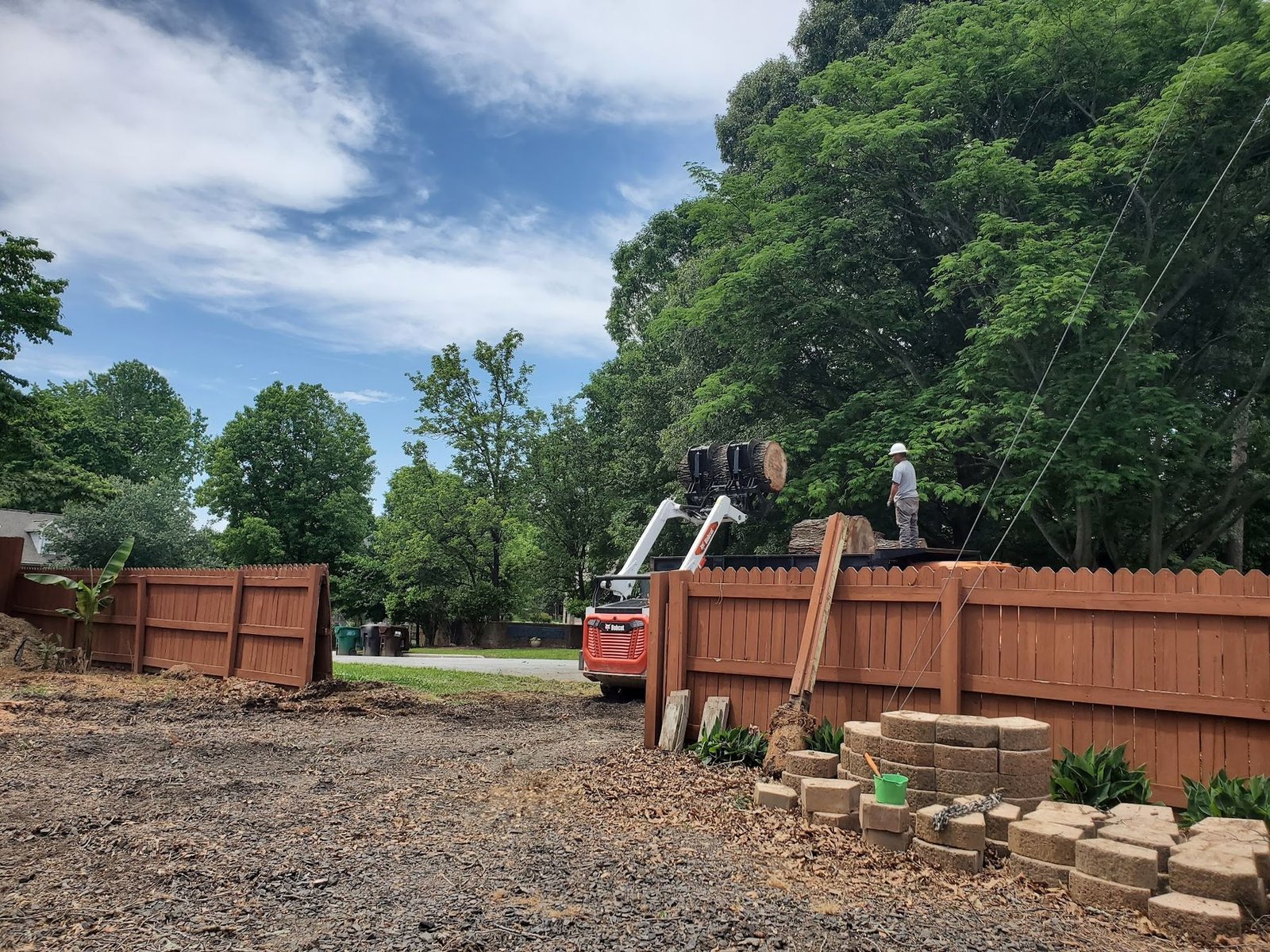 Skid steer lifting a log over a wooden fence into a dump truck on a cleared lot