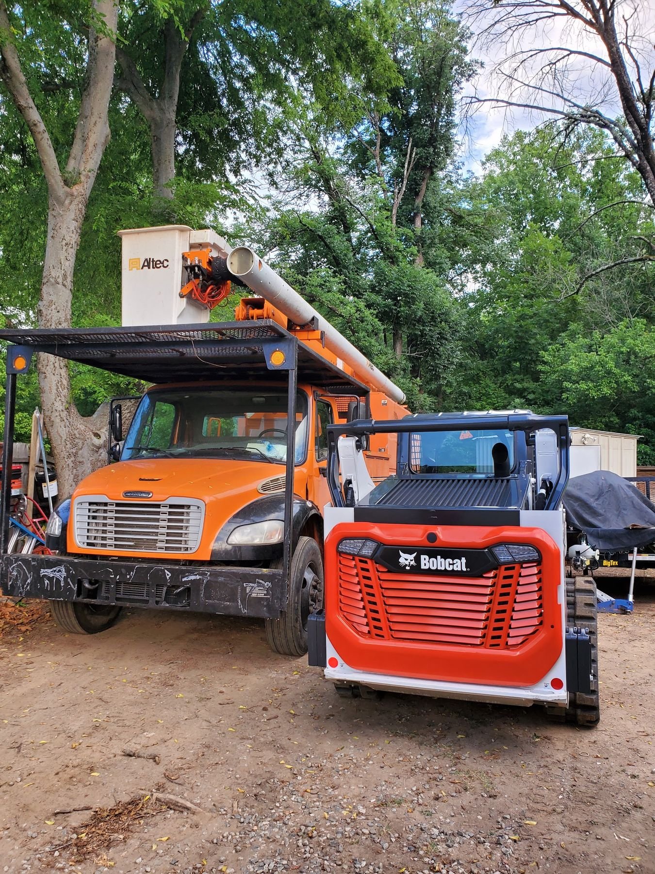 Bucket truck and a new Bobcat track loader parked together in a wooded lot