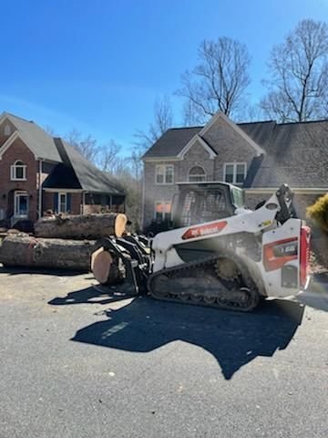 Skid steer grapple carrying a large log round in a driveway in front of a brick home